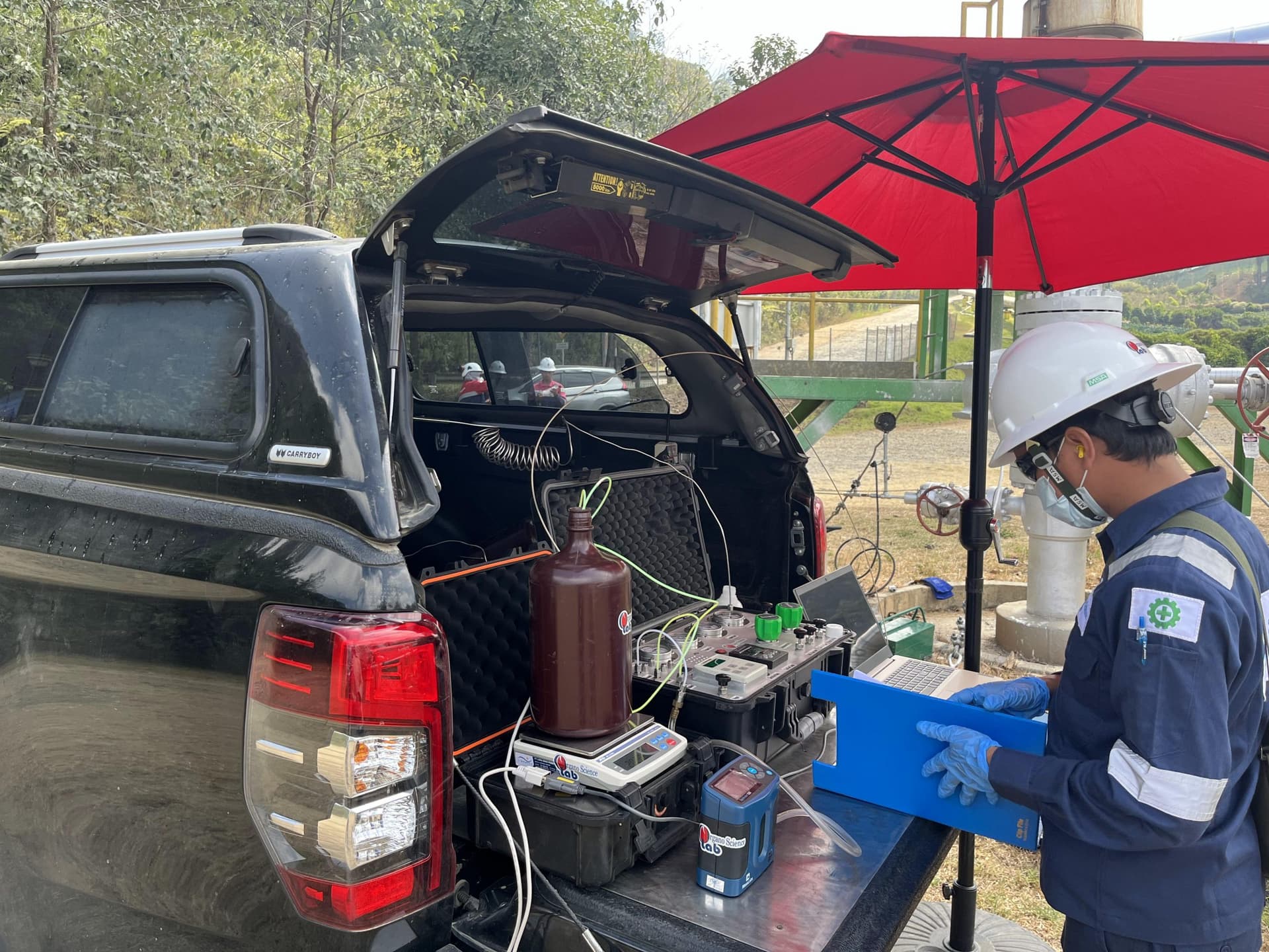 Peralatan sampling dan laptop disusun di bak kendaraan lapangan, seorang teknisi berhelm bekerja di bawah payung.