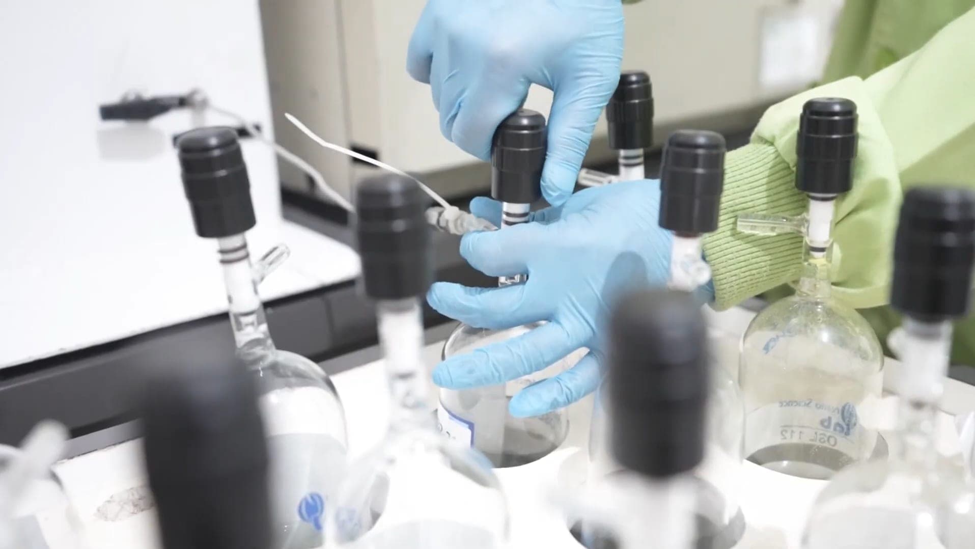 Tangan bersarung merangkai labu kaca bertutup hitam di laboratorium.