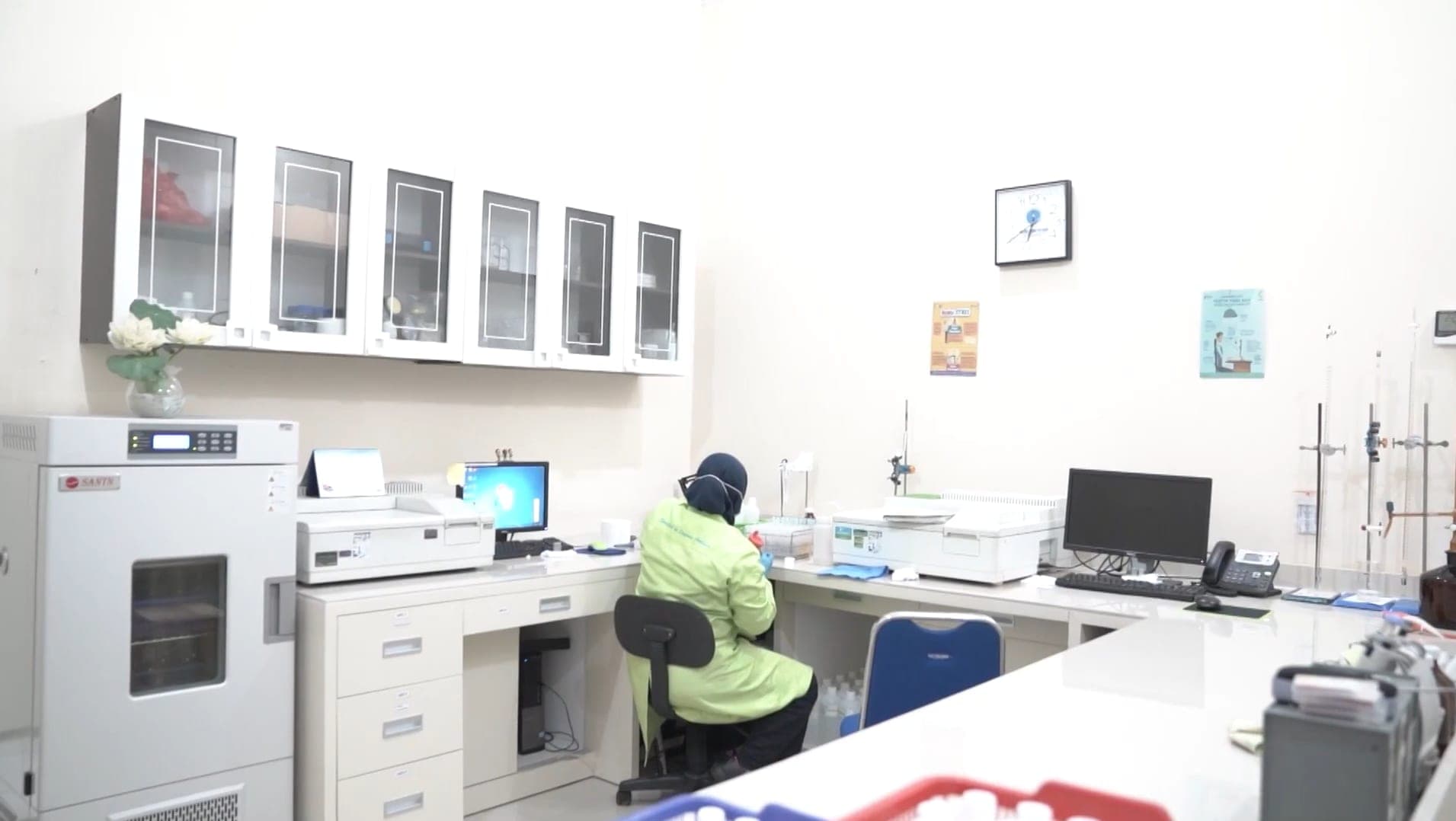 Analis berjas laboratorium hijau bekerja di meja instrumen dalam ruang laboratorium yang terang.