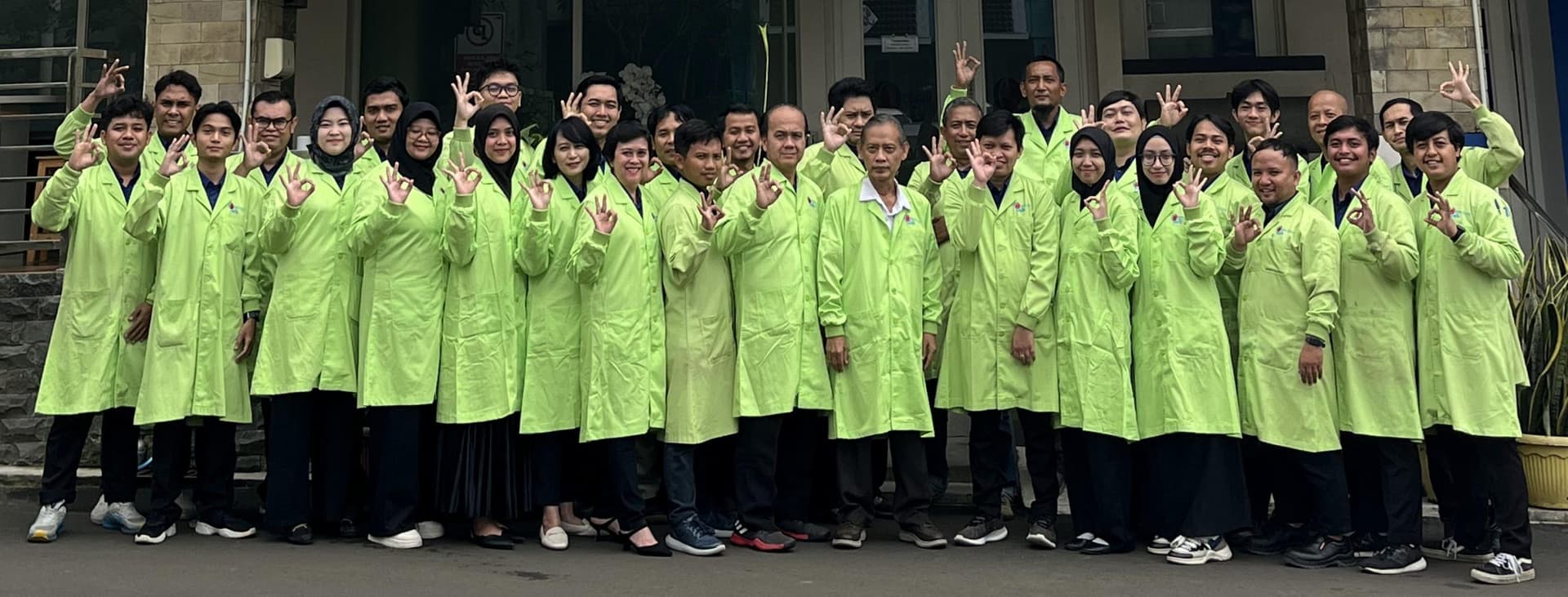 Foto bersama tim berjas laboratorium hijau di depan gedung kantor.