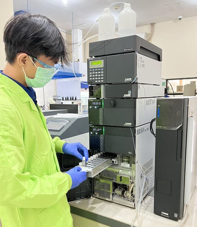 Analis berjas laboratorium hijau memegang rak vial di depan sistem kromatografi cair.